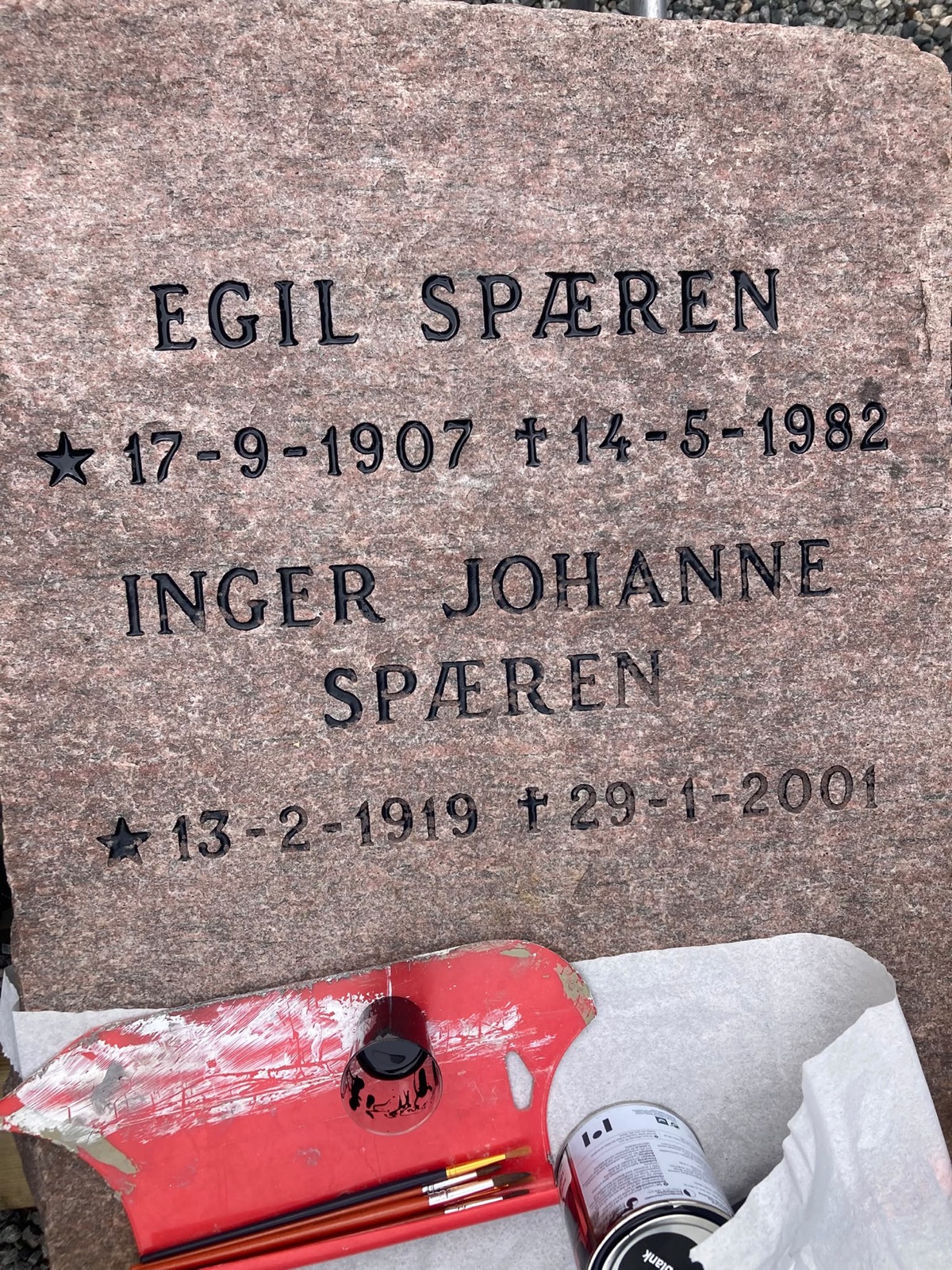 Ferdig oppmalt svart skrift på den rensede, rødlige granitten, med pensler og lakkboks liggende inntil steinen.