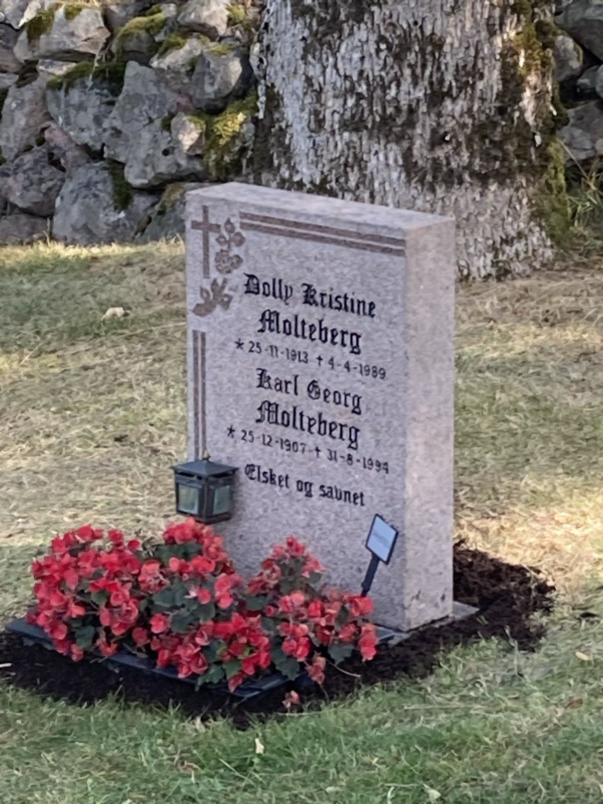 Den ferdige steinen sett på skrå, rett på sokkelen, med røde begonia, lykt og ny jord rundt.