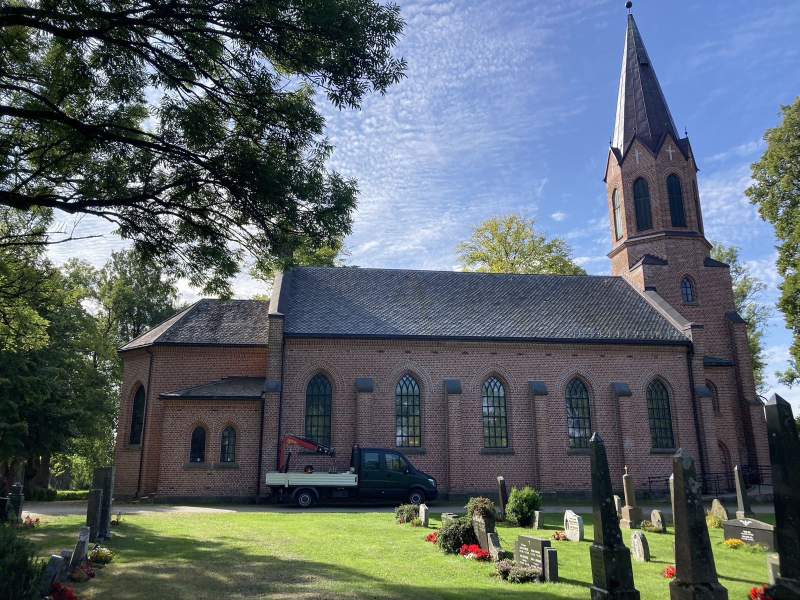 Ås kirke i rød tegl en solrik dag, med arbeidsbilen parkert ved kirkegården.