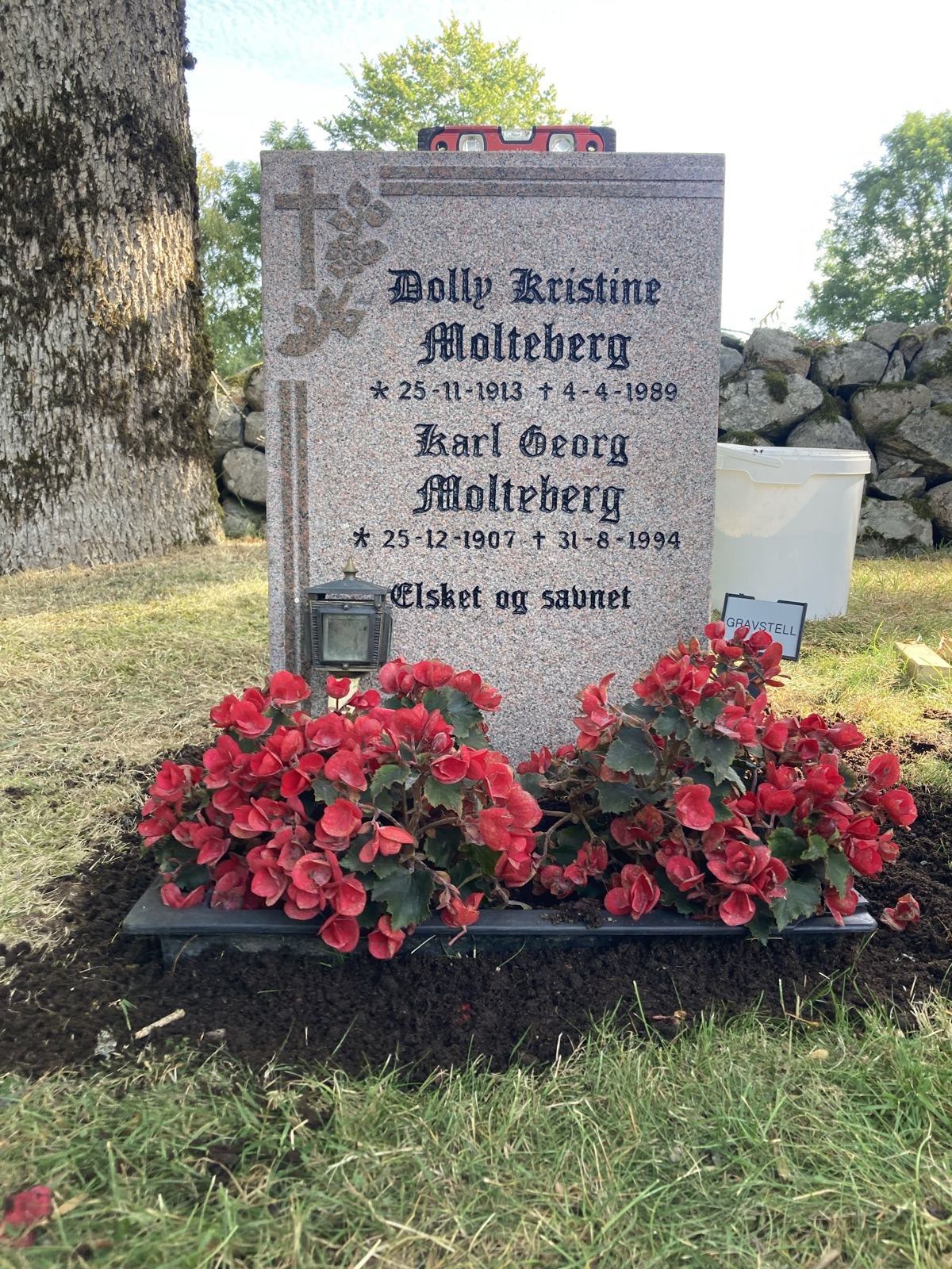 Den ferdige gravsteinen sett forfra med begonia og lykt, foran steinmuren på kirkegården.