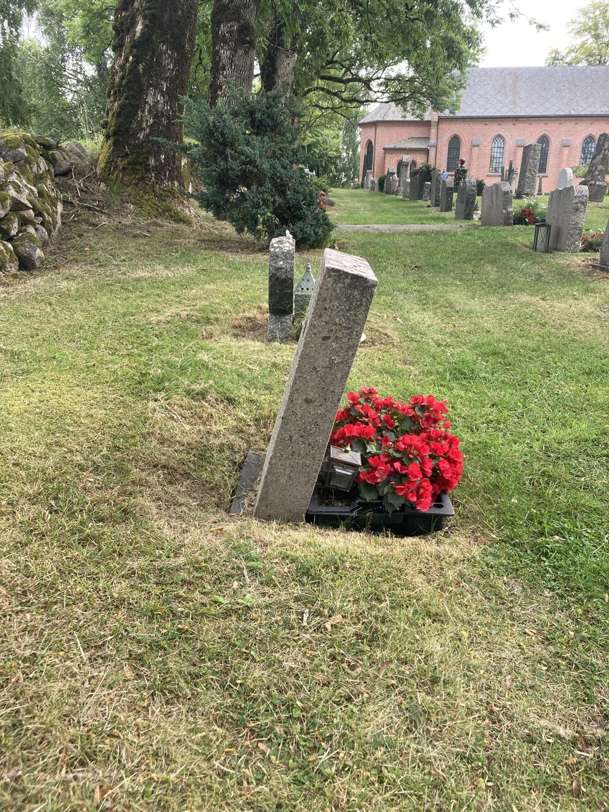 Gravsteinen sett fra siden på kirkegården, hellende forover, med Ås kirke i bakgrunnen.