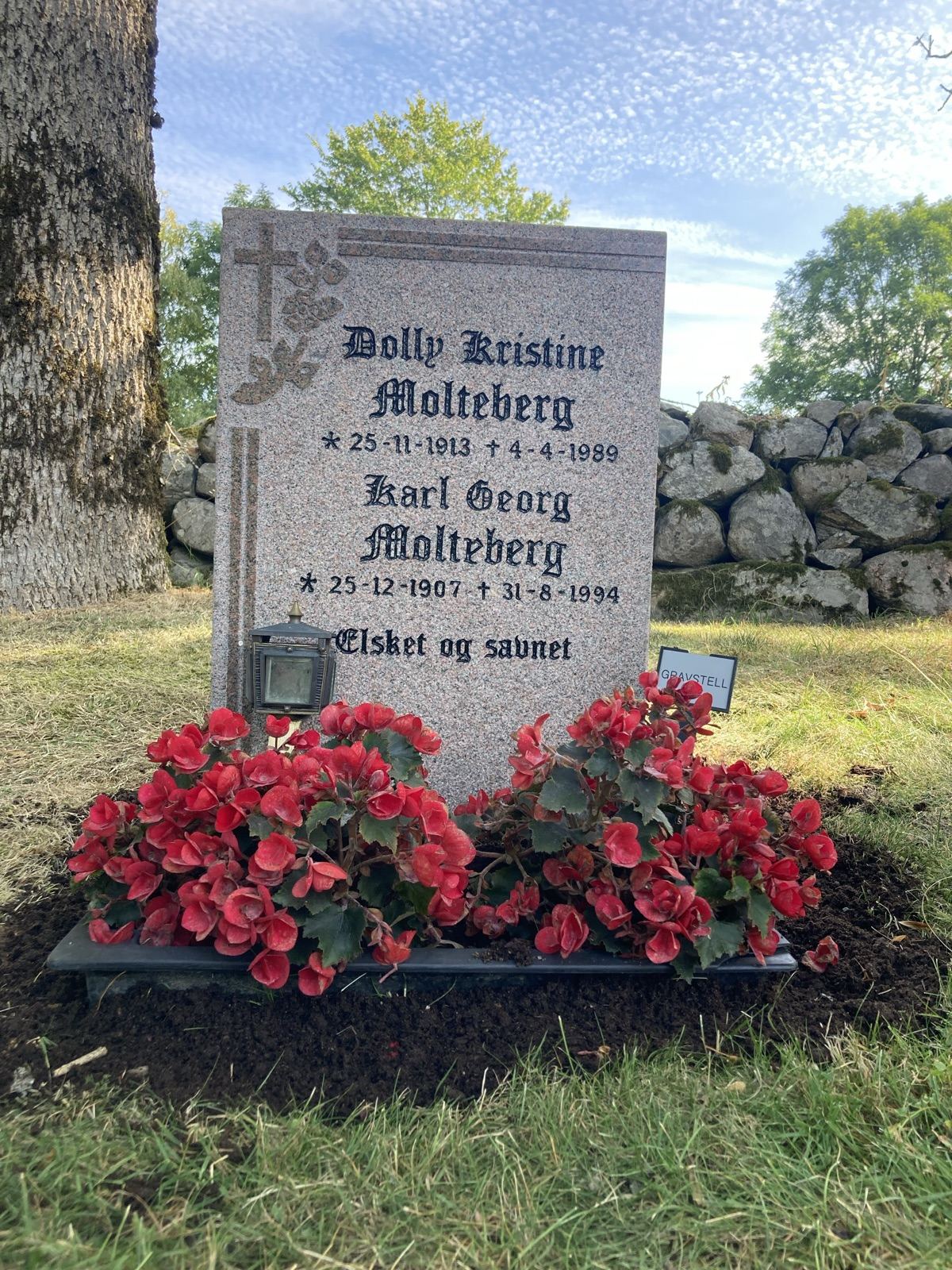 Ferdig pusset gravstein i lys granitt med nymalt mørk skrift, rett på plass foran en steinmur, med røde begonia og en lykt foran.